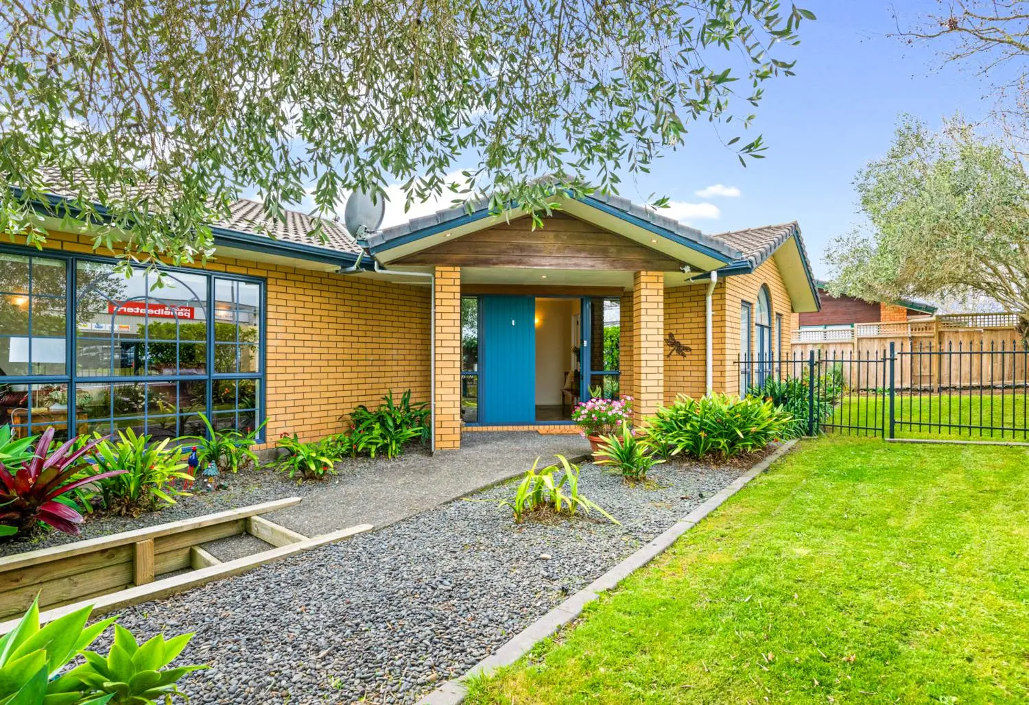 Front entrance of a single-level brick home with a bright blue door, landscaped garden, and neat lawn under mature trees.