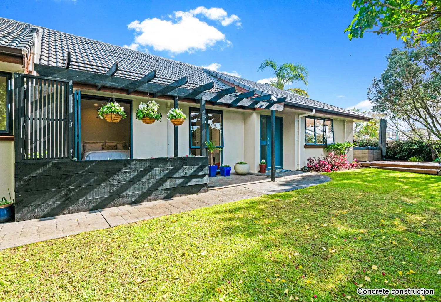 Single-level home with pergola-covered patio, hanging flower baskets, and sunny lawn surrounded by mature trees and gardens.