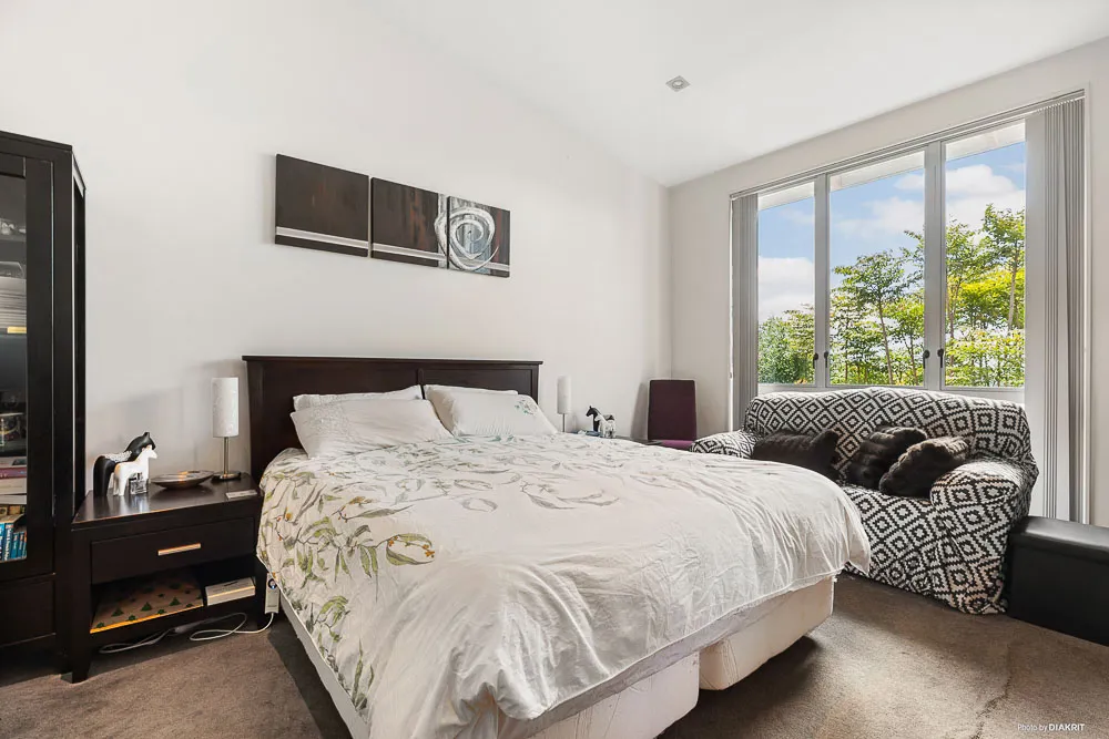 Sunny master bedroom featuring large windows with leafy views, high ceilings, and space for both a queen bed and a cosy sitting area.
